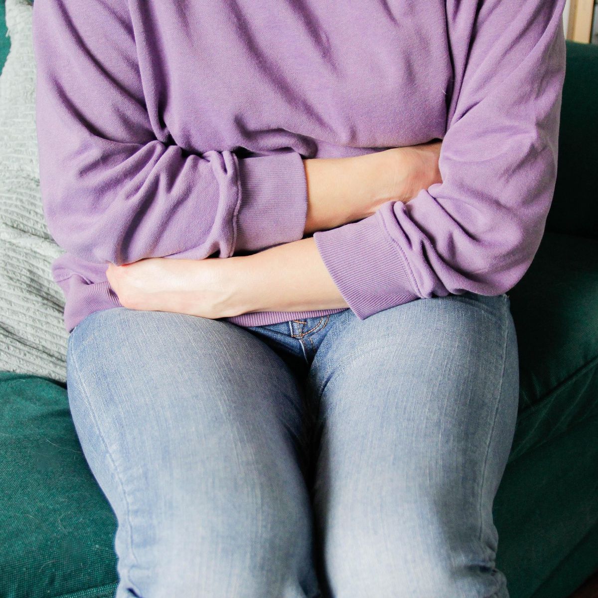 Personne en pull violet assise sur un canapé vert, tenant son ventre avec douleur, indiquant un mal de ventre.