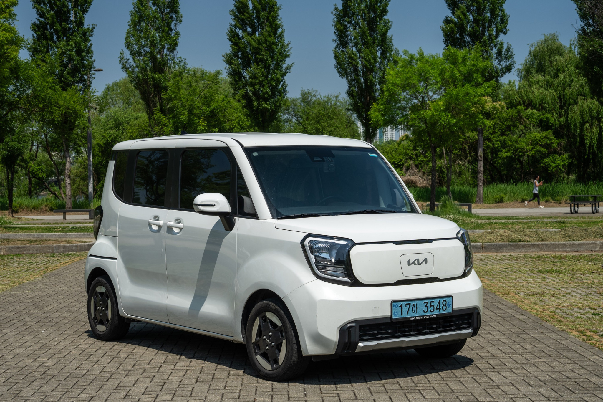 Weisser Kia Ray EV, kompaktes Elektroauto mit geräumigem Innenraum, parkiert auf Pflastersteinen vor grünen Bäumen.
