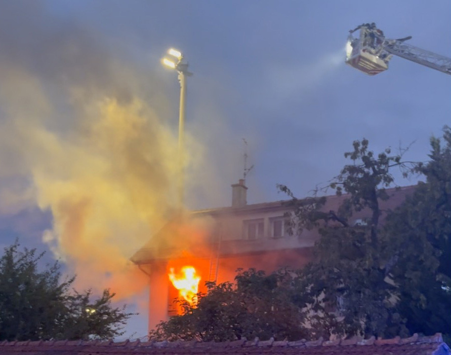 Incendie en cours dans un bâtiment avec de grandes flammes s'échappant d'une fenêtre. Un camion de pompiers avec une échelle est visible en haut à droite, projecteur allumé.