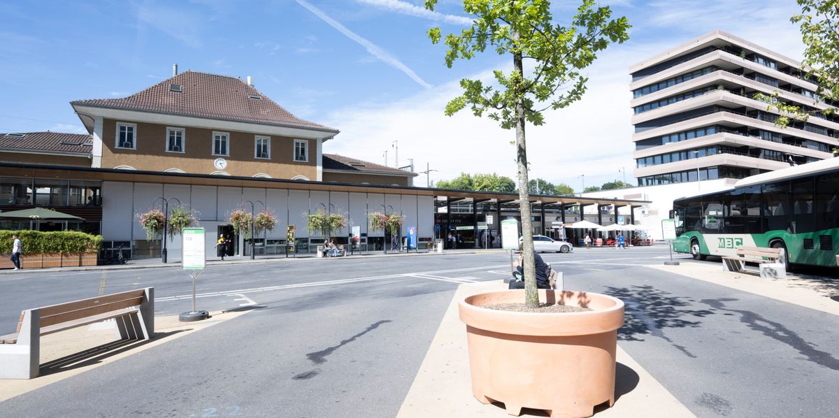 Place de la Gare à Morges avec un bâtiment ferroviaire, un bus vert stationné et un arbre au centre de la place.