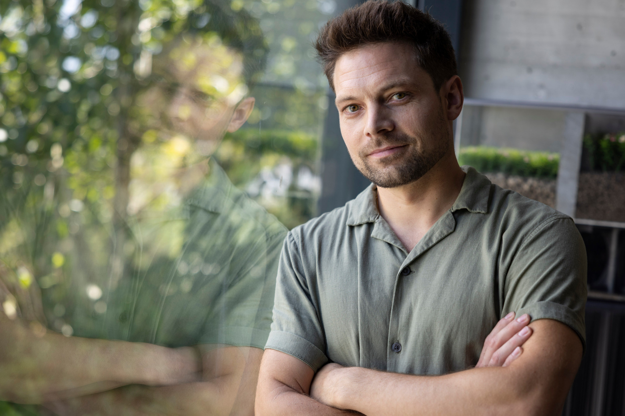 Simon Schopfer steht mit verschränkten Armen vor einem Fenster, das einen grünen Dachgarten reflektiert. Hintergrund zeigt Bäume und Beton.