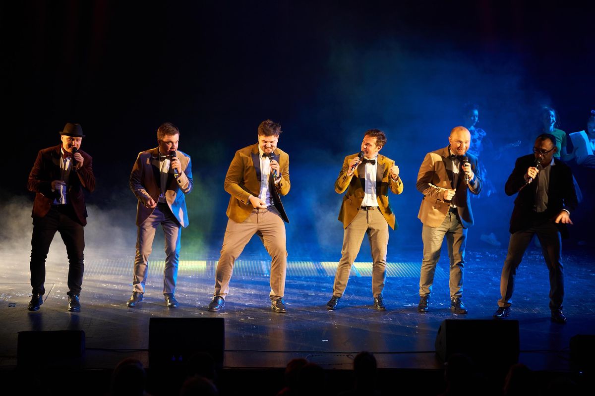 Groupe de six hommes chantant sur scène avec des vestes élégantes, dans une ambiance lumineuse et colorée.