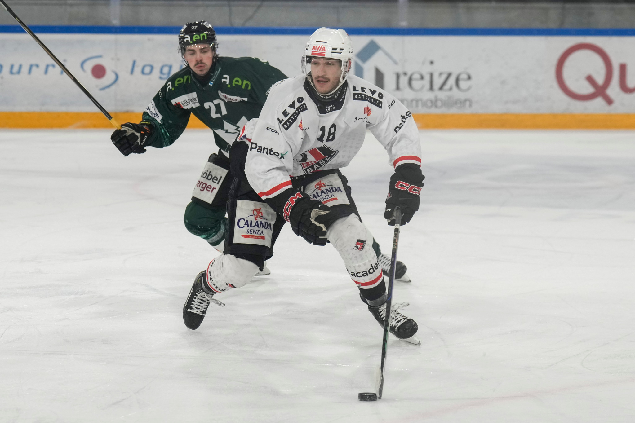 EHC Olten Spieler Devin Stehli im weissen Trikot beim Eishockeyspiel gegen EHC Basel, verfolgt von Lucas Bachofner in grünem Trikot.