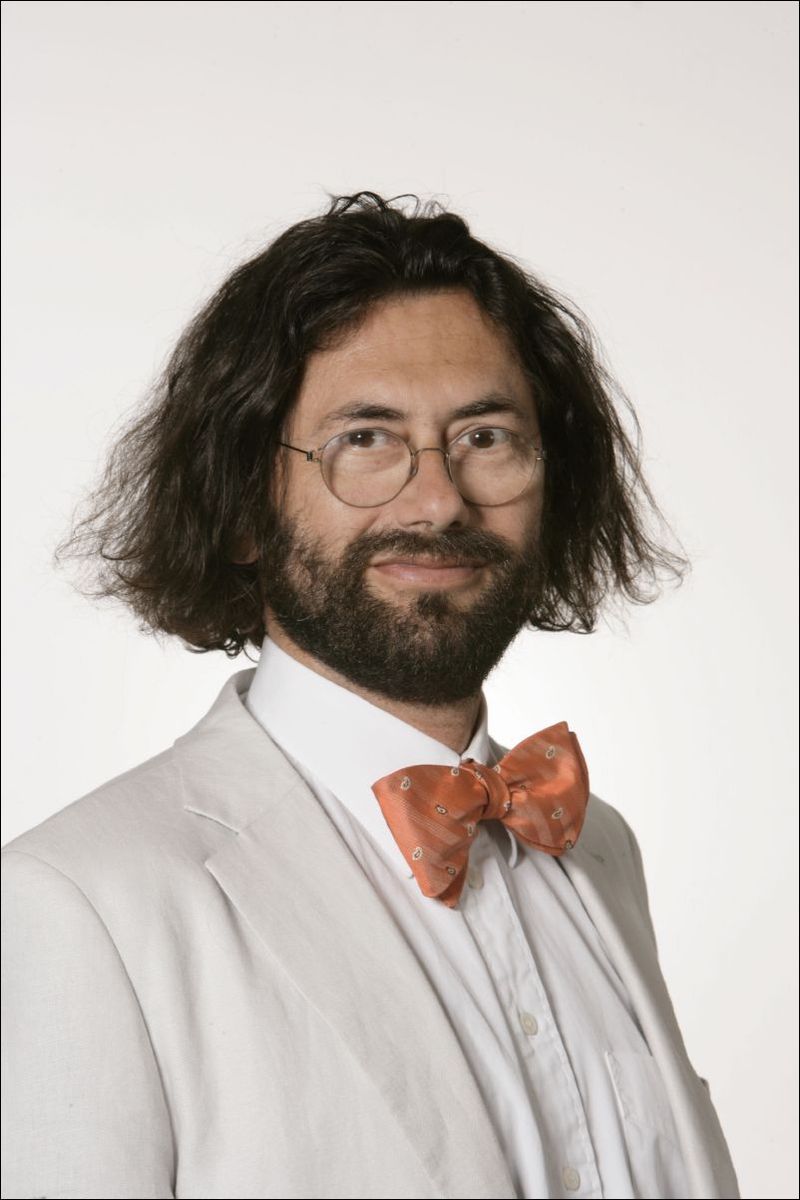 Homme portant un costume blanc avec un nœud papillon orange et des lunettes, sur fond neutre.