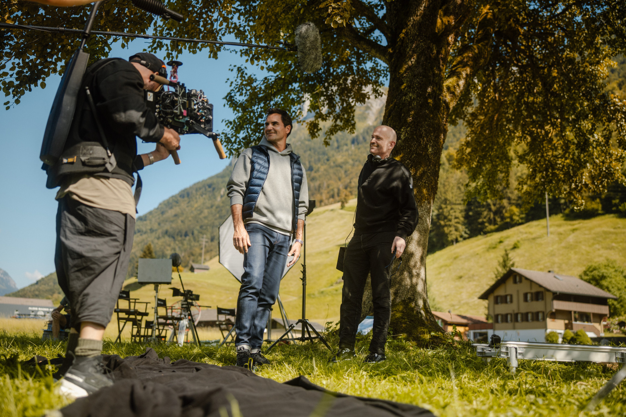 Deux personnes assises à une table en bois sous un parasol, profitant d’une vue panoramique sur un lac et des montagnes par une journée ensoleillée.