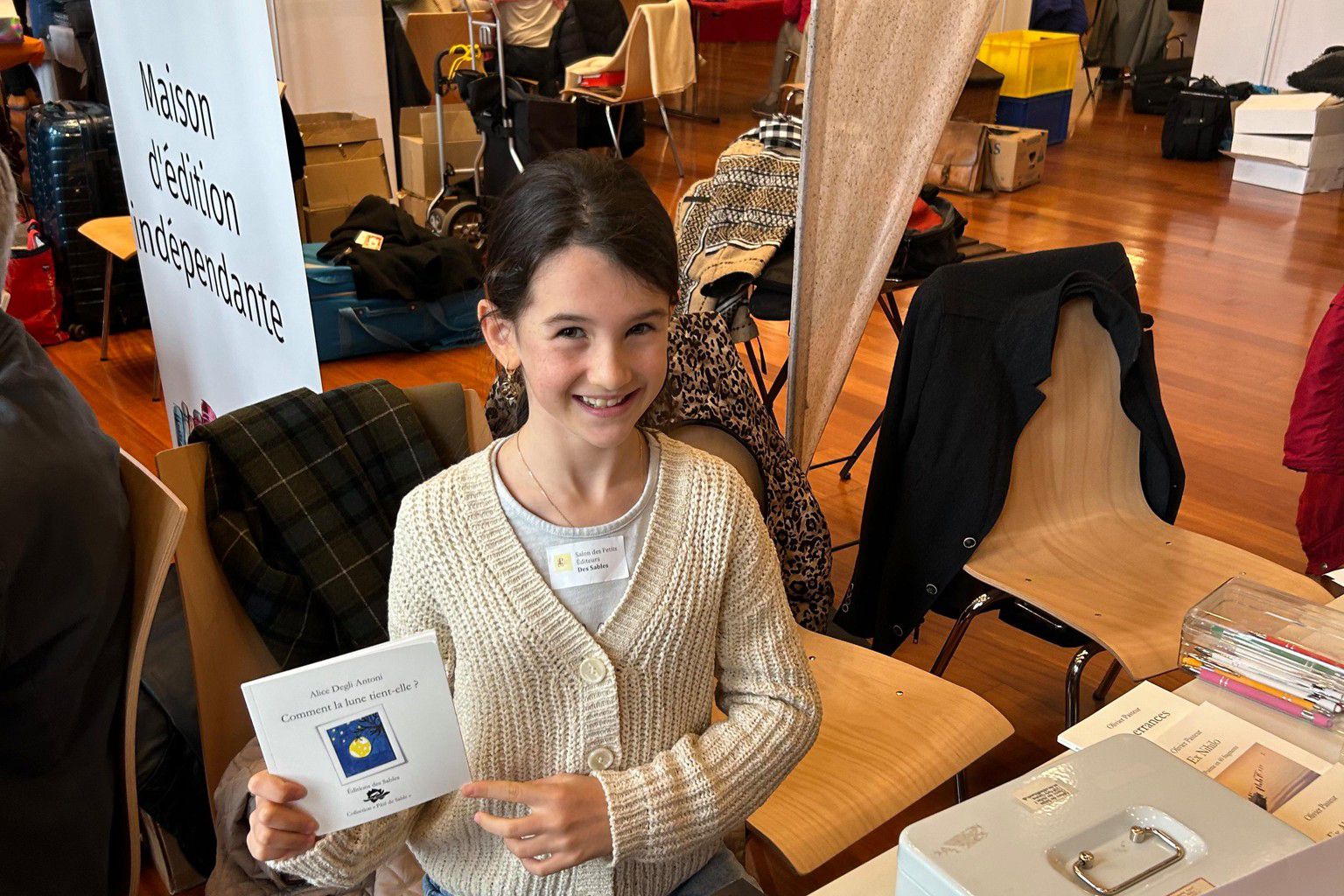 Jeune fille souriante tenant un livre devant un stand de salon littéraire, entourée de tables couvertes de livres.