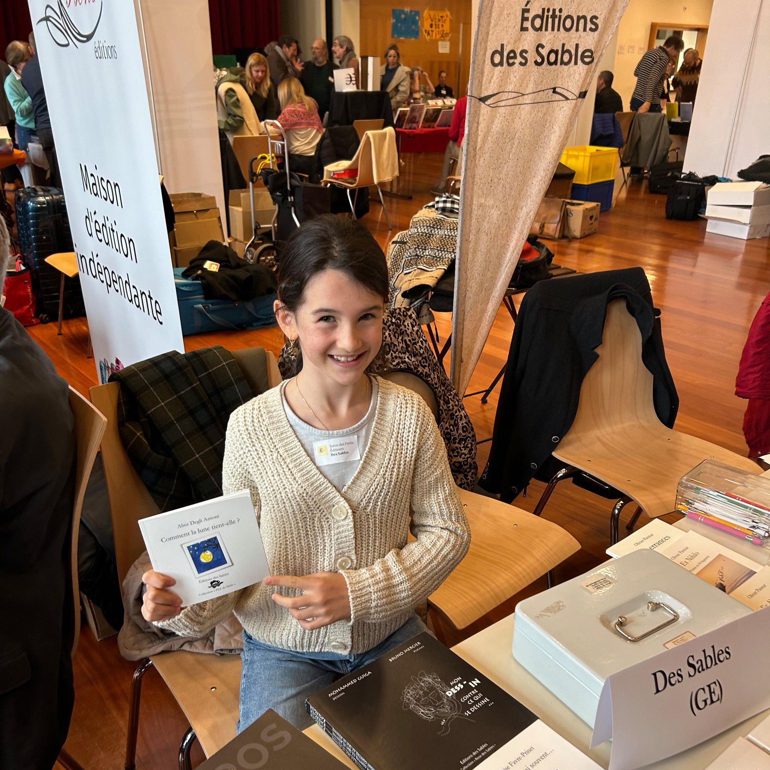 Jeune fille souriante tenant un livre devant un stand de salon littéraire, entourée de tables couvertes de livres.