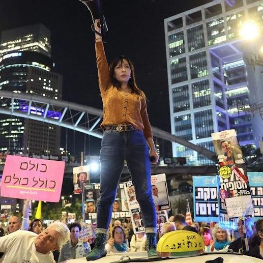 Frau steht auf einem Auto mit Megafon bei einer nächtlichen Demonstration in der Stadt. Umgeben von Menschen mit Plakaten vor beleuchteten Hochhäusern.
