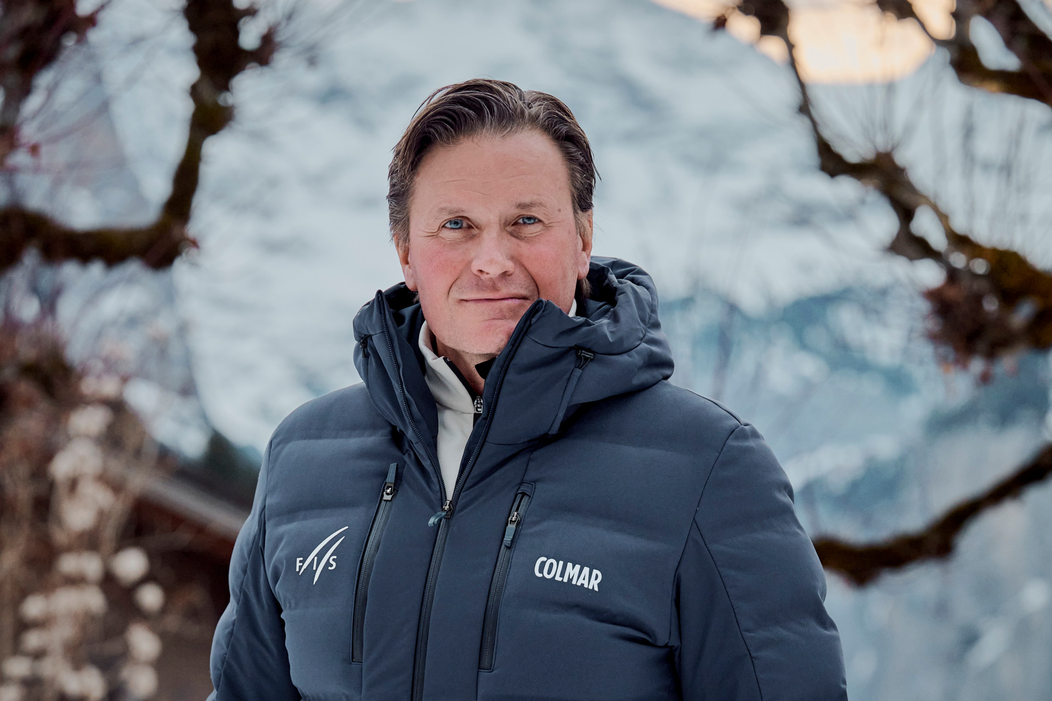 CEO des Internationalen Ski- und Snowboard-Verbandes FIS in blauer Colmar-Jacke mit FIS-Logo vor verschneiter Bergkulisse.