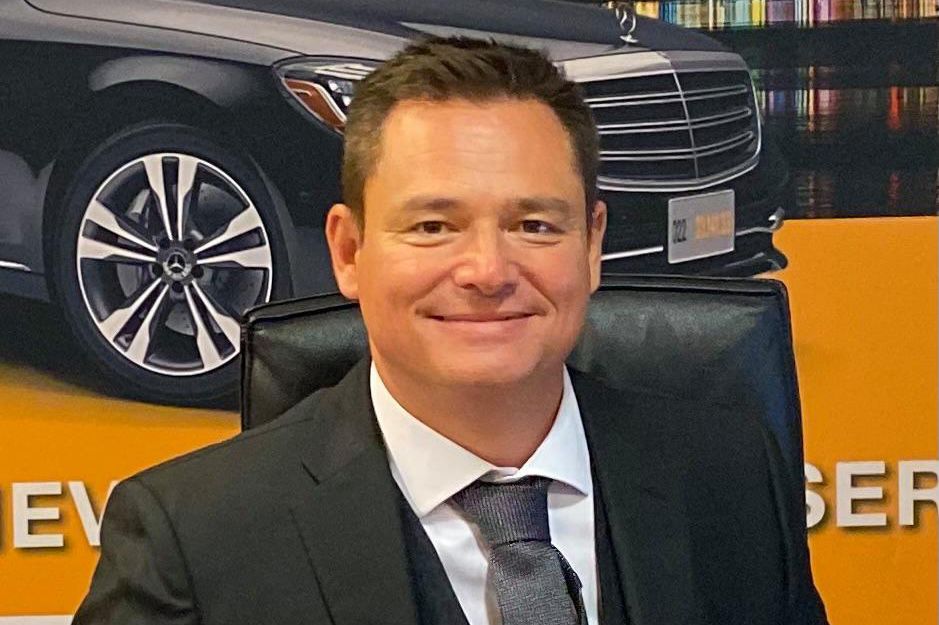 Cédric Bouchard, patron de Taxiphone, en costume assis à un bureau, tenant un stylo, avec une affiche de voiture et vue nocturne en arrière-plan.