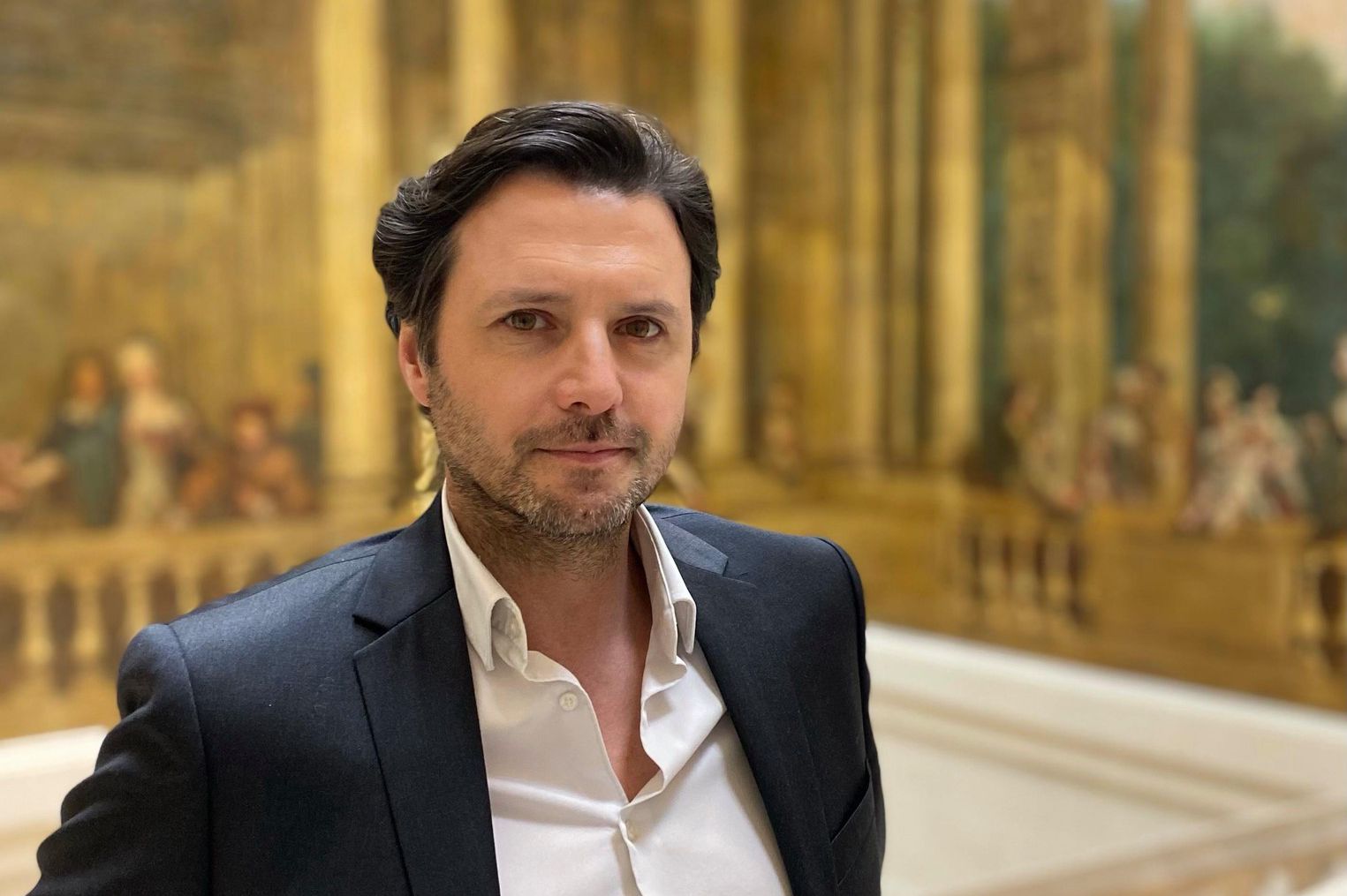 Homme en costume se tenant devant une fresque murale classique dans un bâtiment élégant avec de grandes colonnes.