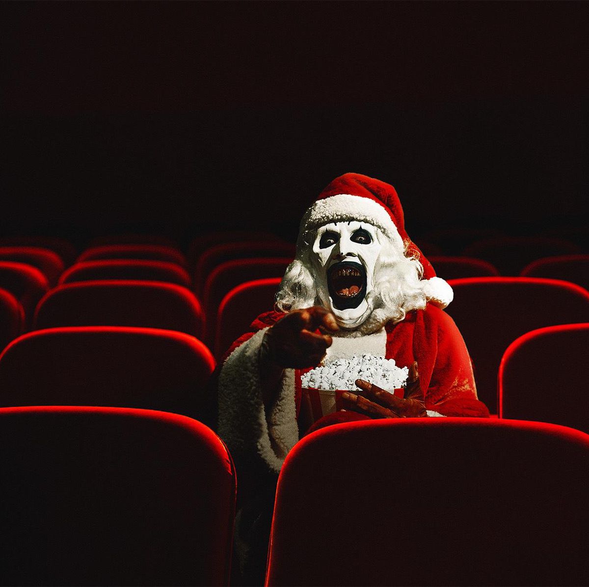 Personne déguisée en Père Noël effrayant, assise seule dans une salle de cinéma vide, tenant du popcorn.