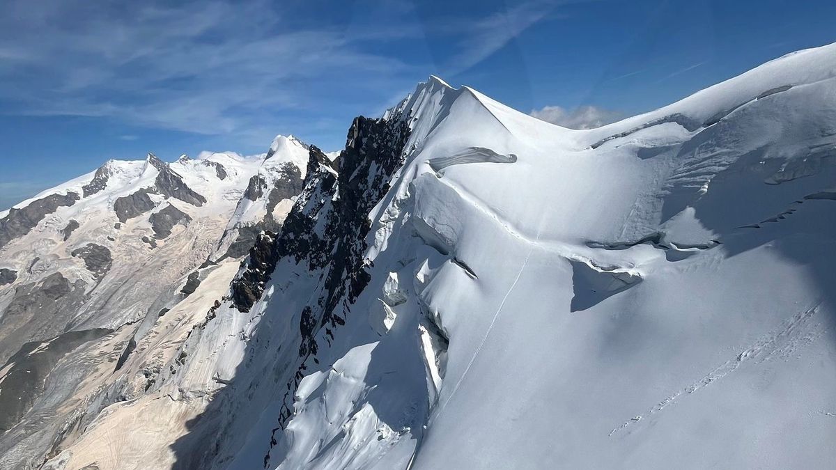 Vue aérienne d'un paysage montagneux avec des pics enneigés et des formations rocheuses sous un ciel bleu clair.