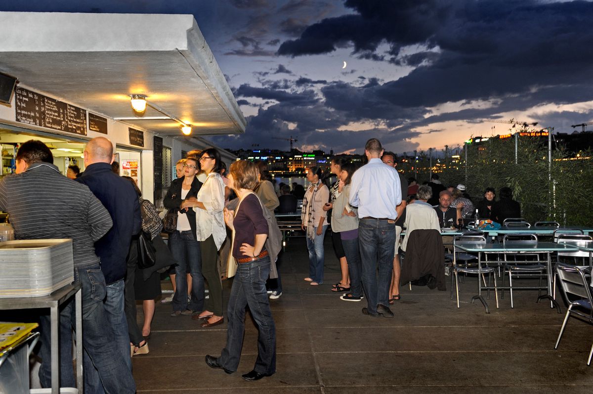 Un groupe de personnes se tient debout et discute près d'un stand de nourriture en plein air en soirée, avec des lumières de la ville et un ciel nuageux en arrière-plan.