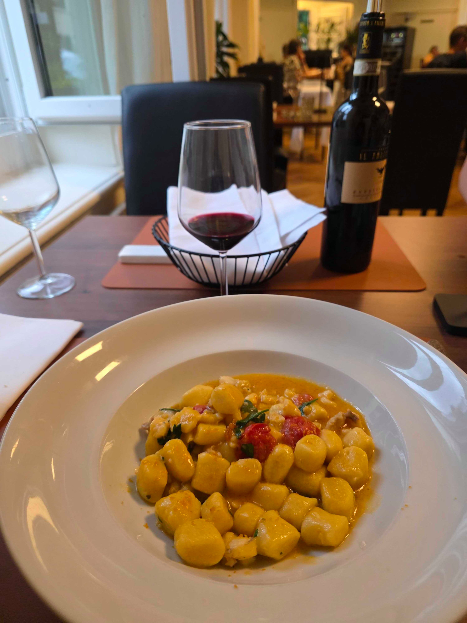 Teller mit Gnocchi in Tomatensauce im Restaurant, im Hintergrund ein Glas Rotwein und eine Weinflasche auf einem Holztisch.