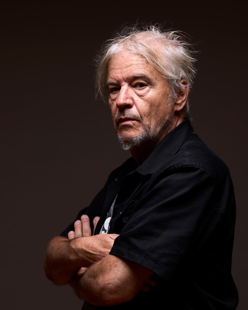Portrait d'un homme âgé avec des cheveux blancs en bataille, portant une chemise noire, avec les bras croisés sur sa poitrine.