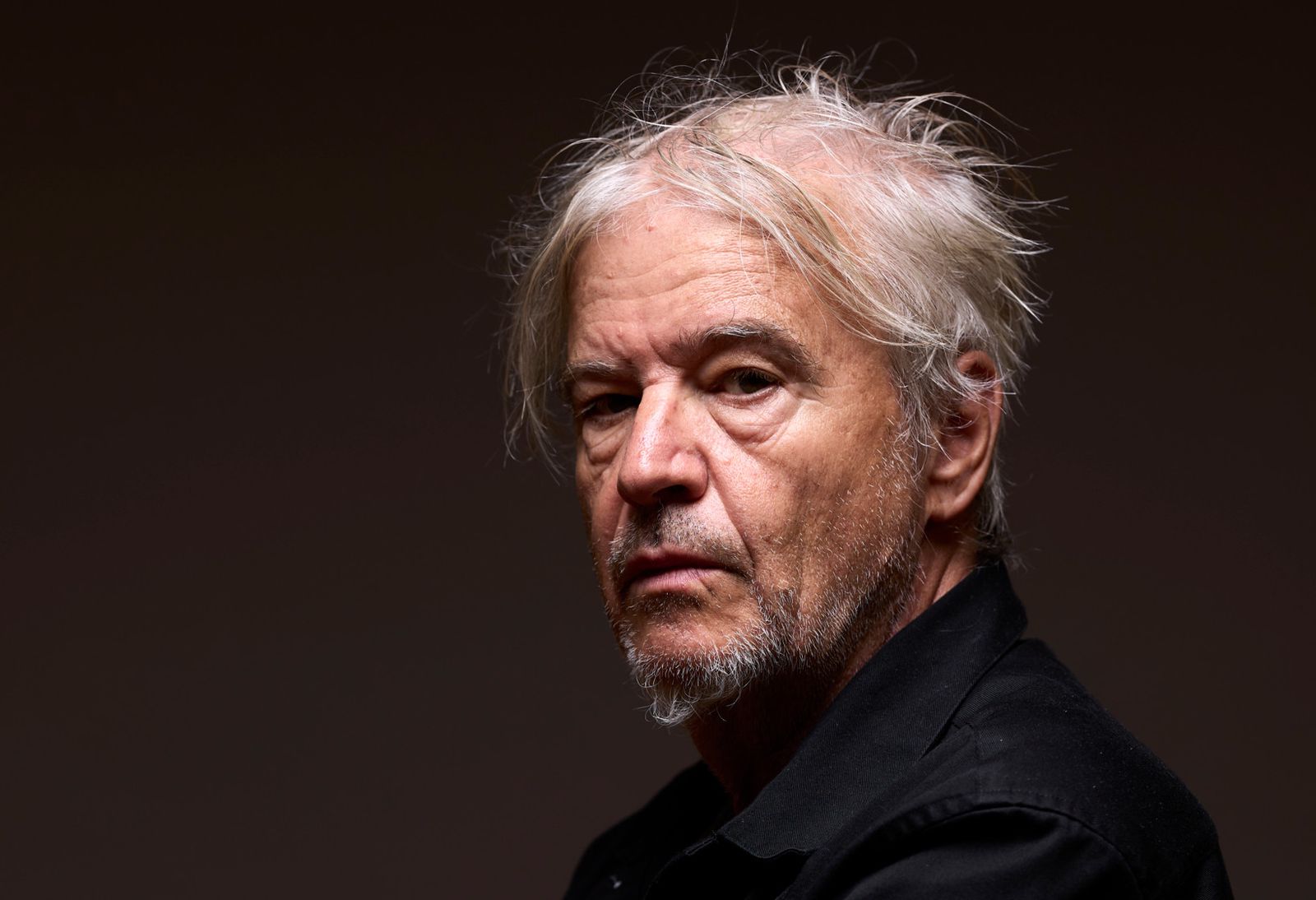 Portrait d'un homme âgé avec des cheveux blancs en bataille, portant une chemise noire, avec les bras croisés sur sa poitrine.
