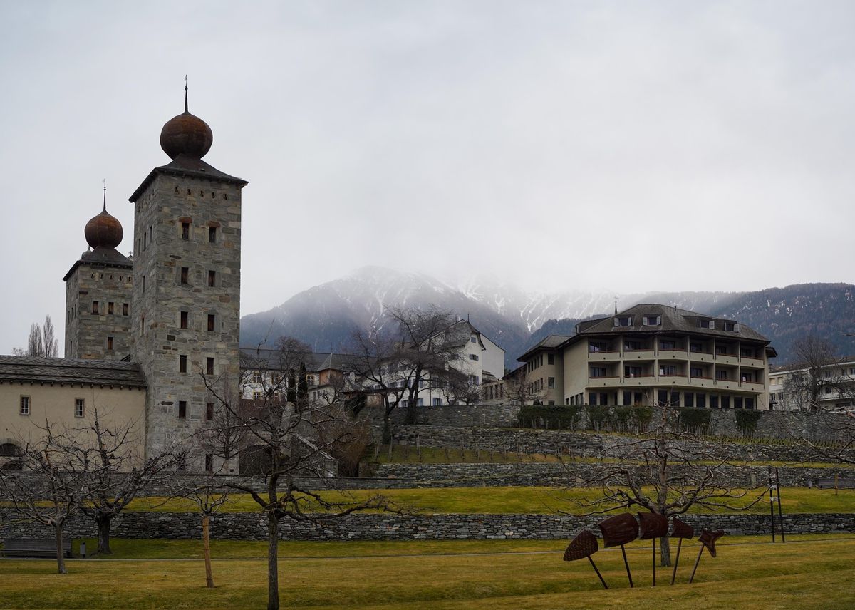 Le bâtiment est situé en plein coeur de la vieille ville de Brigue (maison blanche aux volets bleus et l’immeuble gris adjacent), jouxtant le château Stockalper.