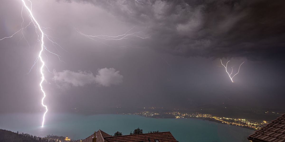 Blitze entladen sich über einem See und einer Stadt bei Nacht, dramatische Wolkenbildung, erleuchtete Küste im Hintergrund.