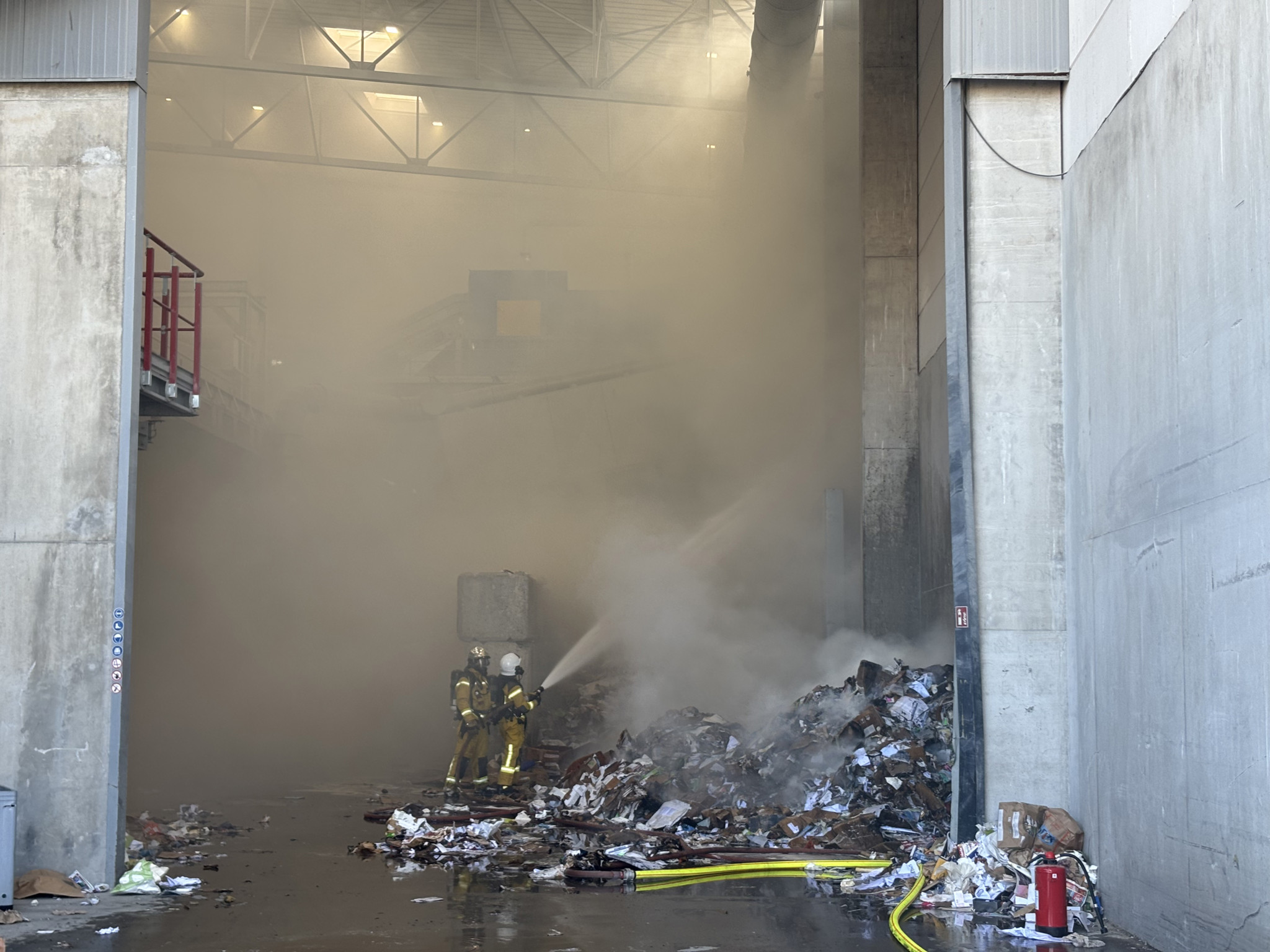 Pompiers luttant contre un incendie dans un bâtiment industriel rempli de fumée, avec des déchets au sol.