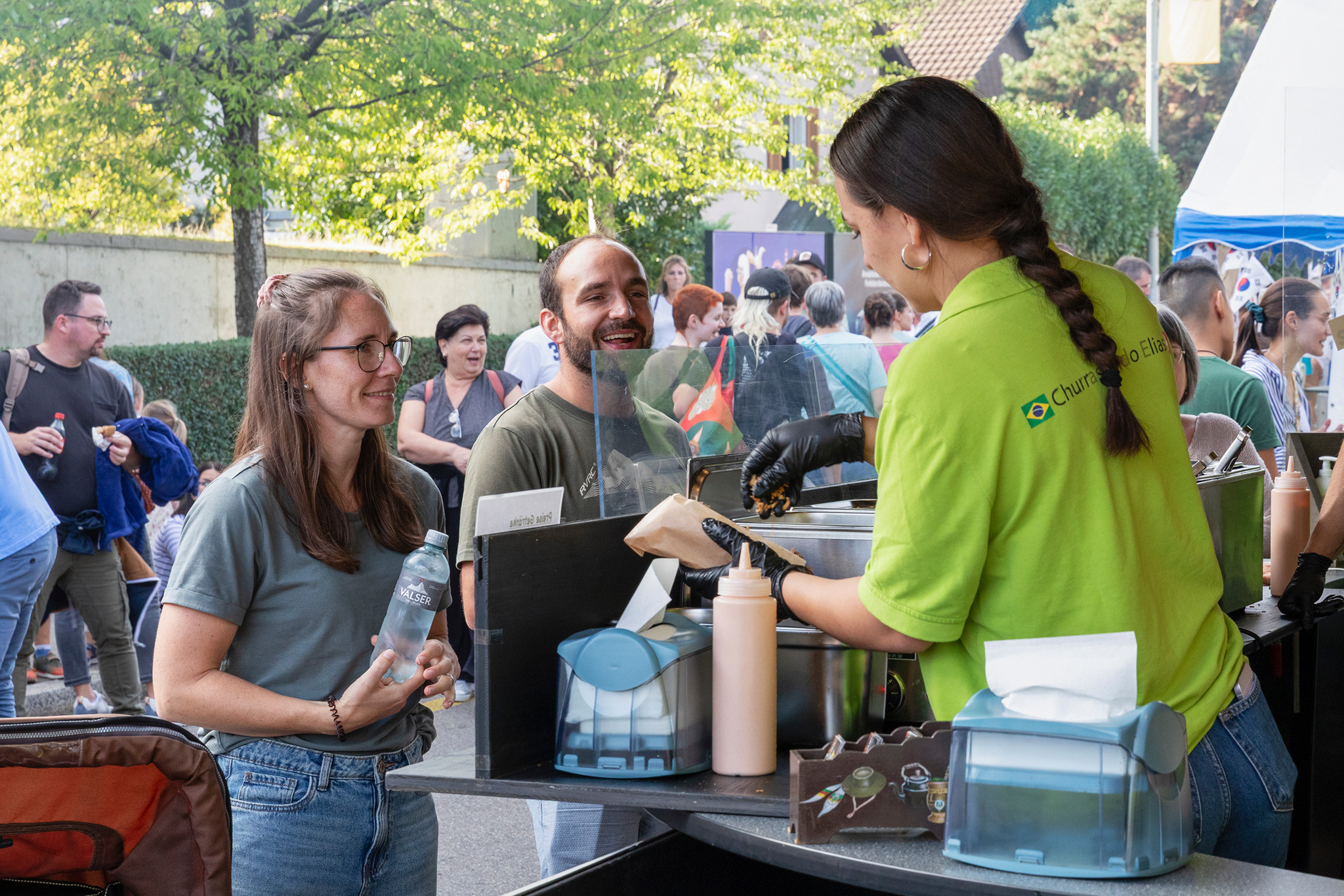 Frau im grünen T-Shirt bedient lächelnde Besucher an einem Foodstand bei einem belebten Outdoor-Festival.