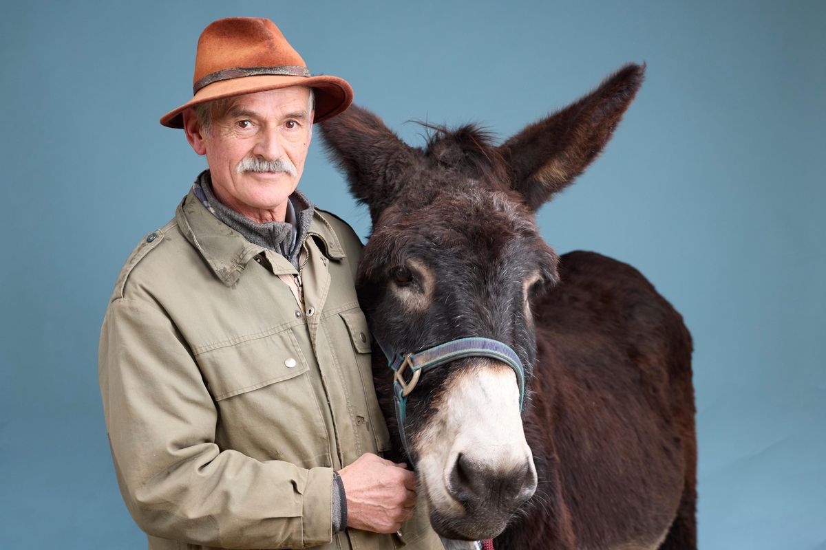 Jean-Michel pose avec son âne Igord, portant un chapeau marron, sur un fond bleu à Cheseaux.