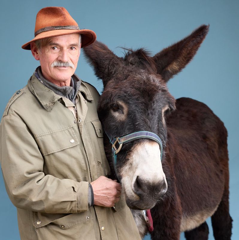 Jean-Michel pose avec son âne Igord, portant un chapeau marron, sur un fond bleu à Cheseaux.