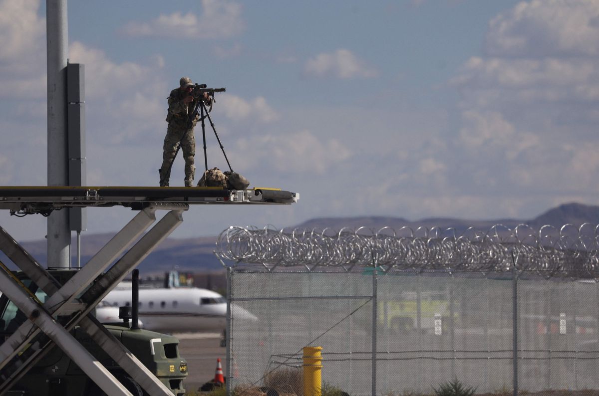 Un soldat en uniforme, posté sur une plateforme surélevée, utilise un viseur pour surveiller une zone entourée de barbelés, avec un avion et des montagnes en arrière-plan.
