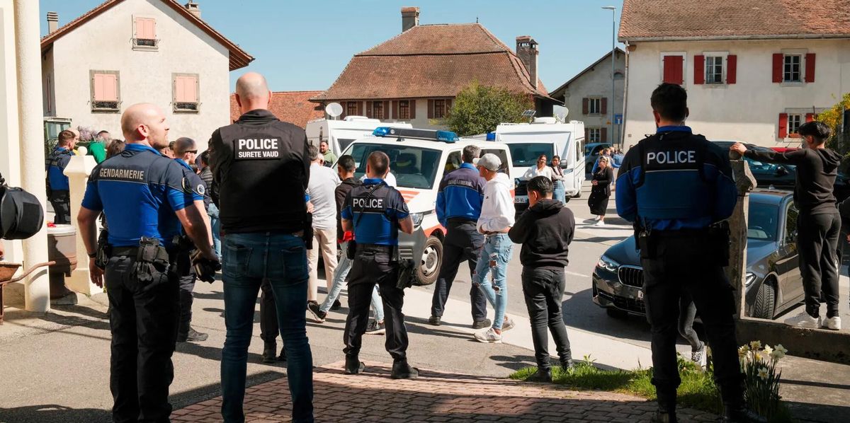 Des policiers en uniforme surveillent un rassemblement de personnes devant des maisons dans une zone résidentielle. Un véhicule de la police est stationné à proximité.
