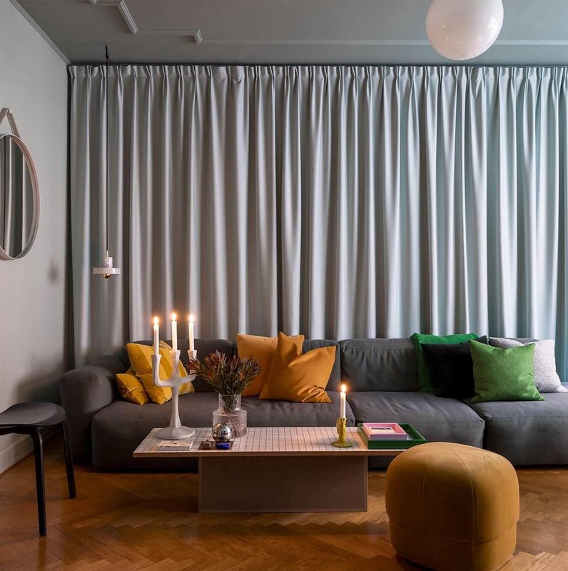 Modernes Wohnzimmer mit grauem Sofa und farbigen Kissen, Couchtisch mit Kerzen, einem Hocker und Vorhängen im Hintergrund.