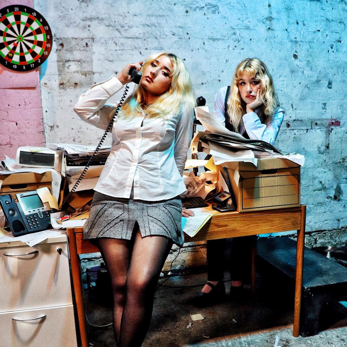 Deux femmes dans un bureau en désordre, l’une au téléphone devant un bureau avec des papiers empilés, l’autre appuyée sur une table. Un jeu de fléchettes est accroché au mur en brique.