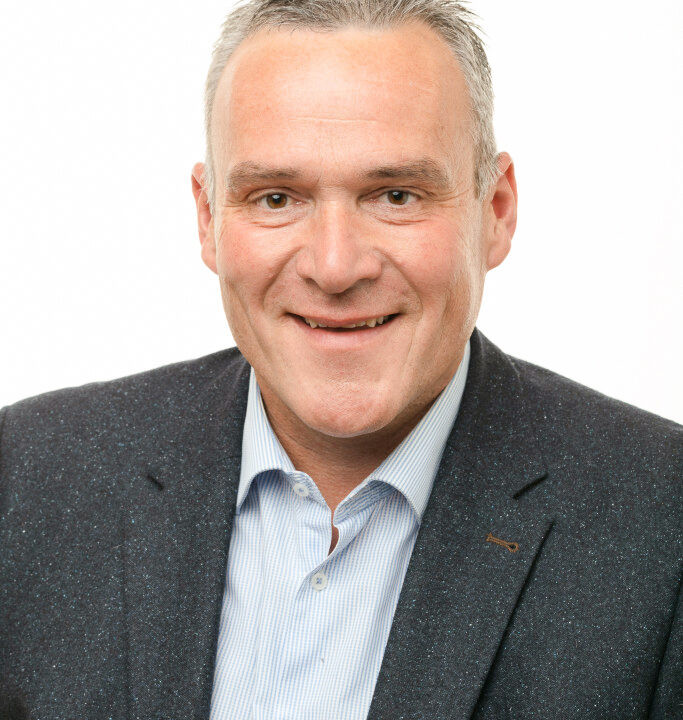 Homme souriant en costume sombre et chemise bleue, regardant directement l’appareil photo.