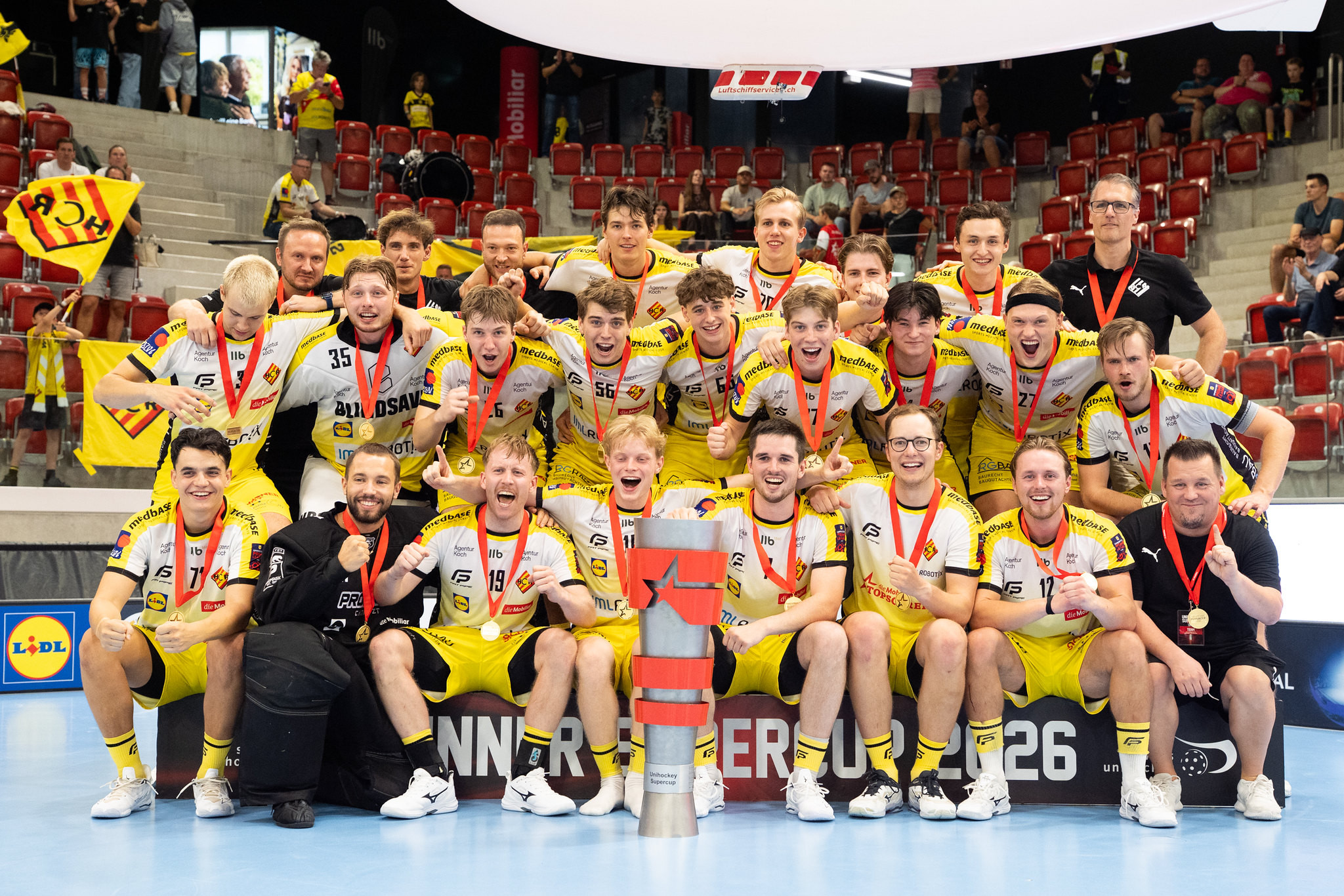 Handballteam in gelb-weissen Trikots feiert mit Medaillen und Pokal den Meistertitel in einer Sporthalle.