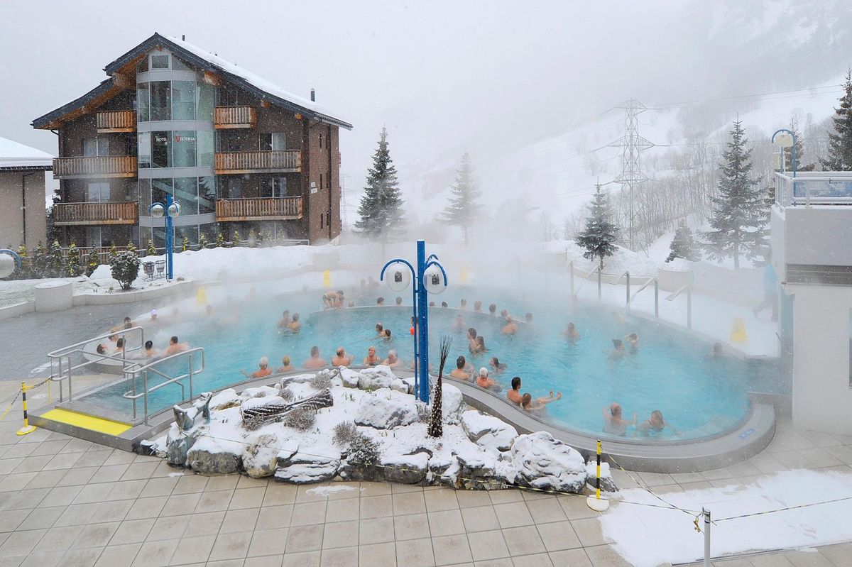 Des personnes se baignent dans une piscine thermale entourée de neige à Loèche-les-Bains, devant un bâtiment alpin.