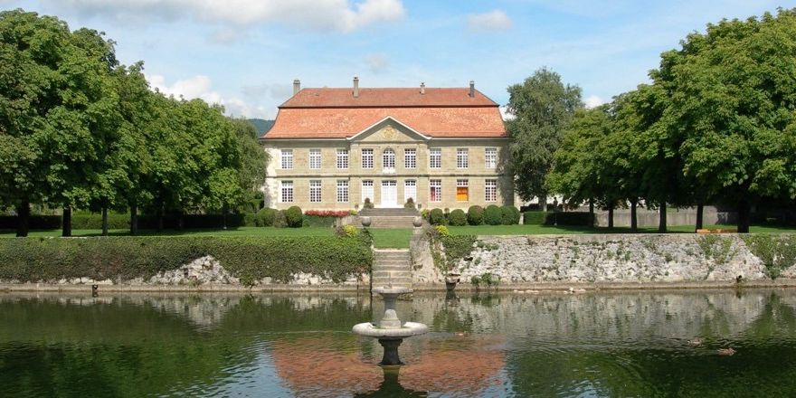 Manoir historique avec un toit rouge entouré d'arbres, avec un étang et une fontaine au premier plan.