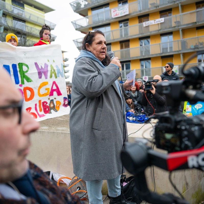 Frau hält bei einer Protestveranstaltung Mikrofon, umgeben von Menschen und Transparenten. Eine Kamera filmt die Szene.
