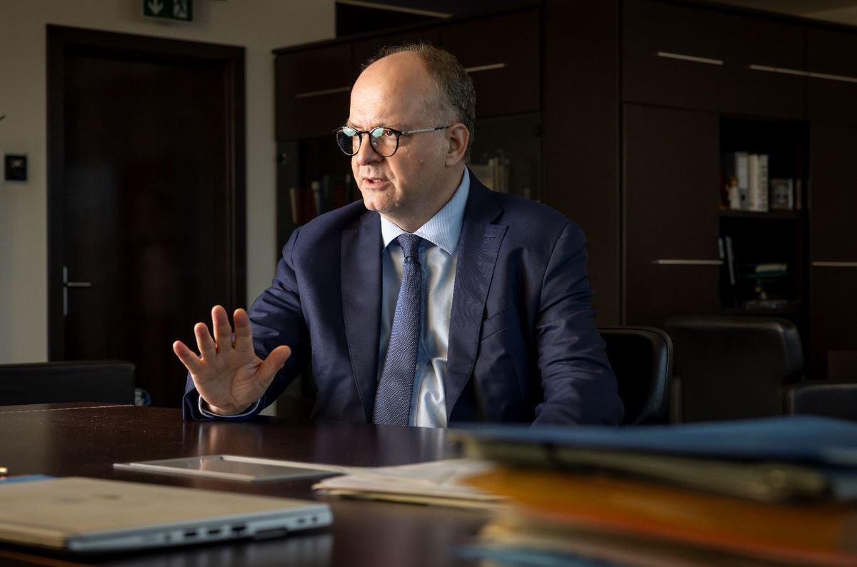 Homme en costume assis à un bureau, main levée, devant des dossiers et un ordinateur portable dans un bureau.