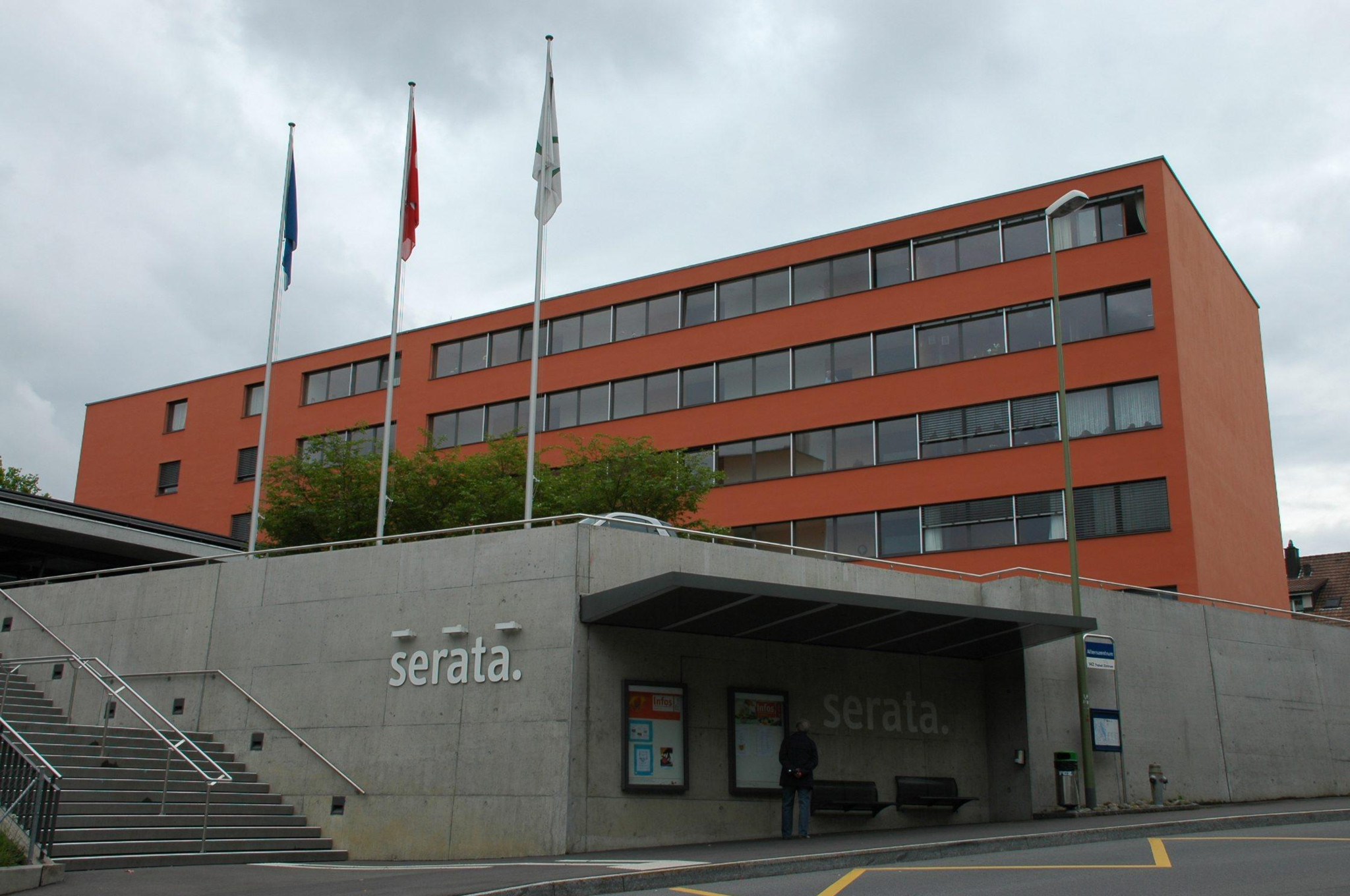 Aussenansicht des Serata-Gebäudes mit orangefassade, drei Fahnenmasten und überdachtem Eingangsbereich bei bewölktem Himmel.