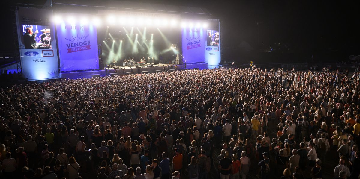 Grande foule assistant à un concert en plein air la nuit, avec une scène illuminée et des écrans géants affichant des performances artistiques.