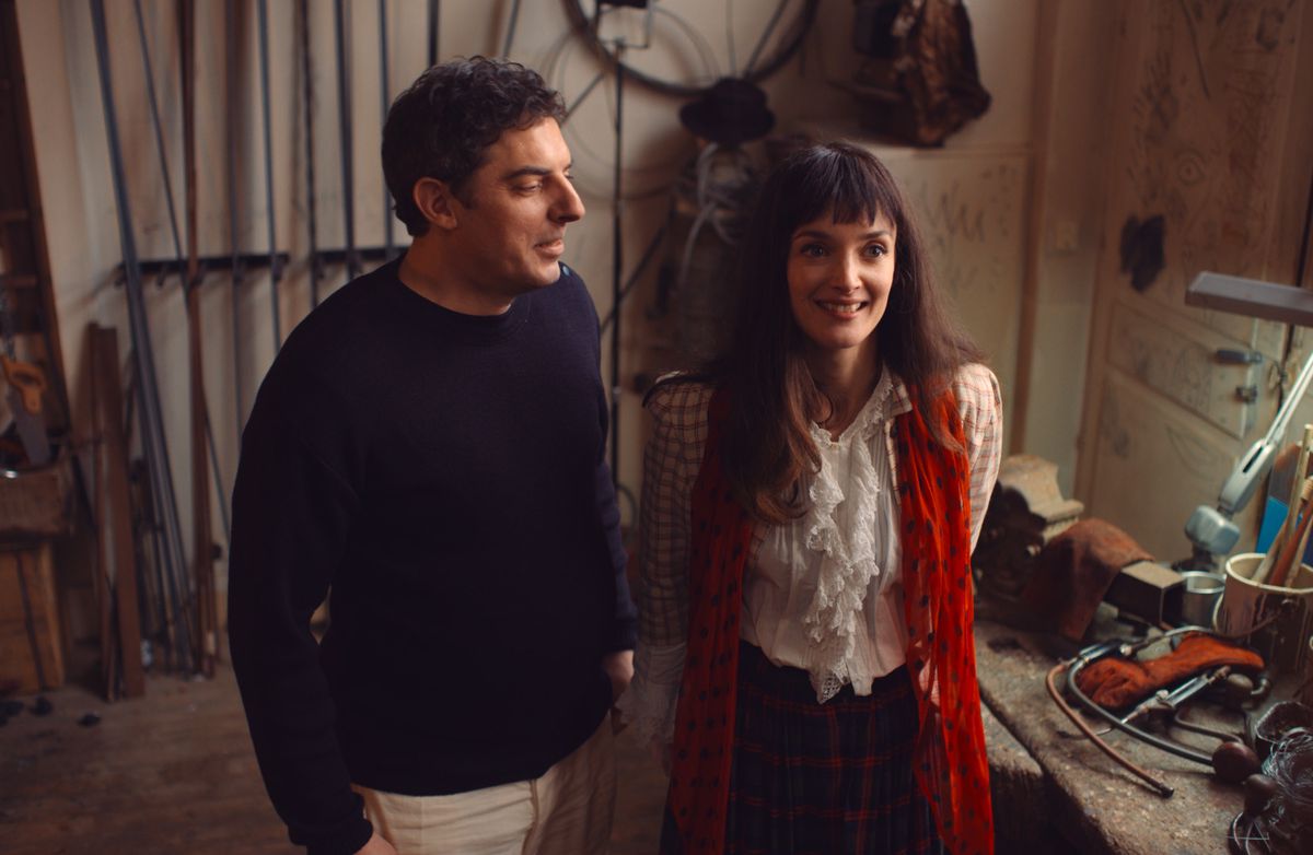 Un homme et une femme souriante debout dans un atelier encombré, avec des outils sur un plan de travail.