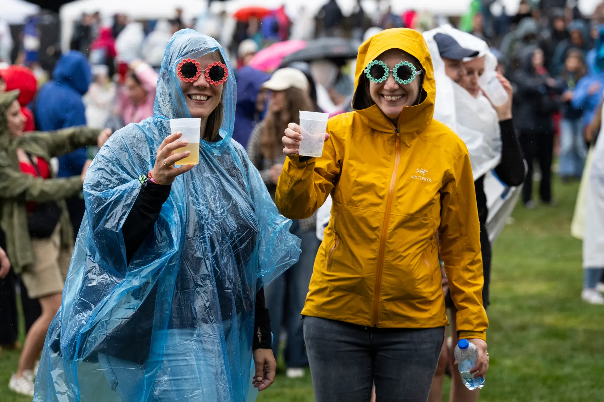 Zwei lachende Frauen mit bunten Blumenbrillen halten Becher auf einem Outdoor-Festival im Regen – eine im blauen Regenponcho, die andere in gelber Jacke. Zwei lachende Frauen mit bunten Blumenbrillen halten Becher auf einem Outdoor-Festival im Regen – eine im blauen Regenponcho, die andere in gelber Jacke.