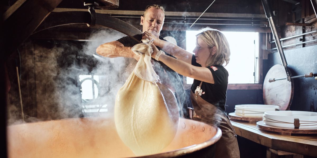 Deux personnes travaillent dans une fromagerie, tenant un sac de fromage chaud au-dessus d'une grande cuve en cuivre.