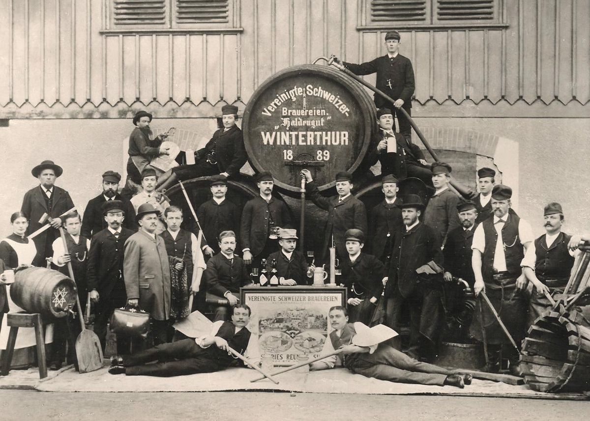 Historische Aufnahme der Arbeiter der Brauerei Haldengut in Winterthur aus dem Jahr 1889. Mehrere Männer posieren vor einem grossen Bierfass mit der Aufschrift ’Vereinigte Schweizer Brauereien Haldengut Winterthur’.