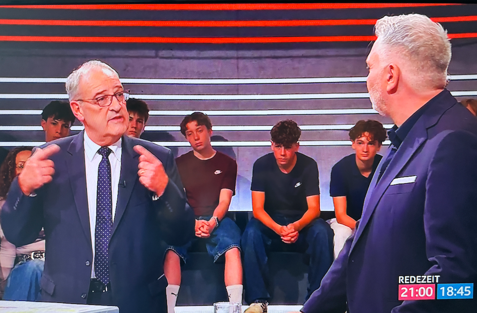 Zwei Männer in Anzügen diskutieren in einer TV-Sendung namens ’Redezeit’, im Hintergrund sitzen Jugendliche als Studiopublikum.