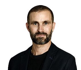 Un homme avec une barbe et des cheveux courts portant une veste noire, posant sur un fond blanc.