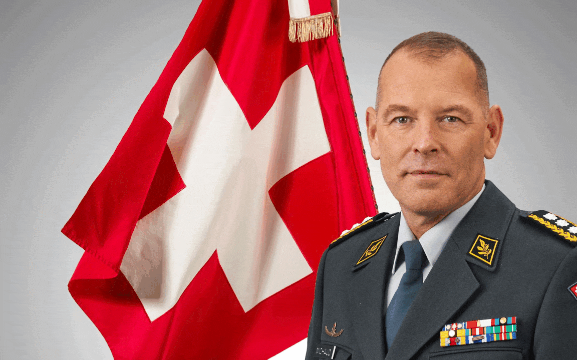 Ein Mann in einer Schweizer Militäruniform sitzt an einem Tisch in einem Raum mit einer Schweizer Fahne im Hintergrund.