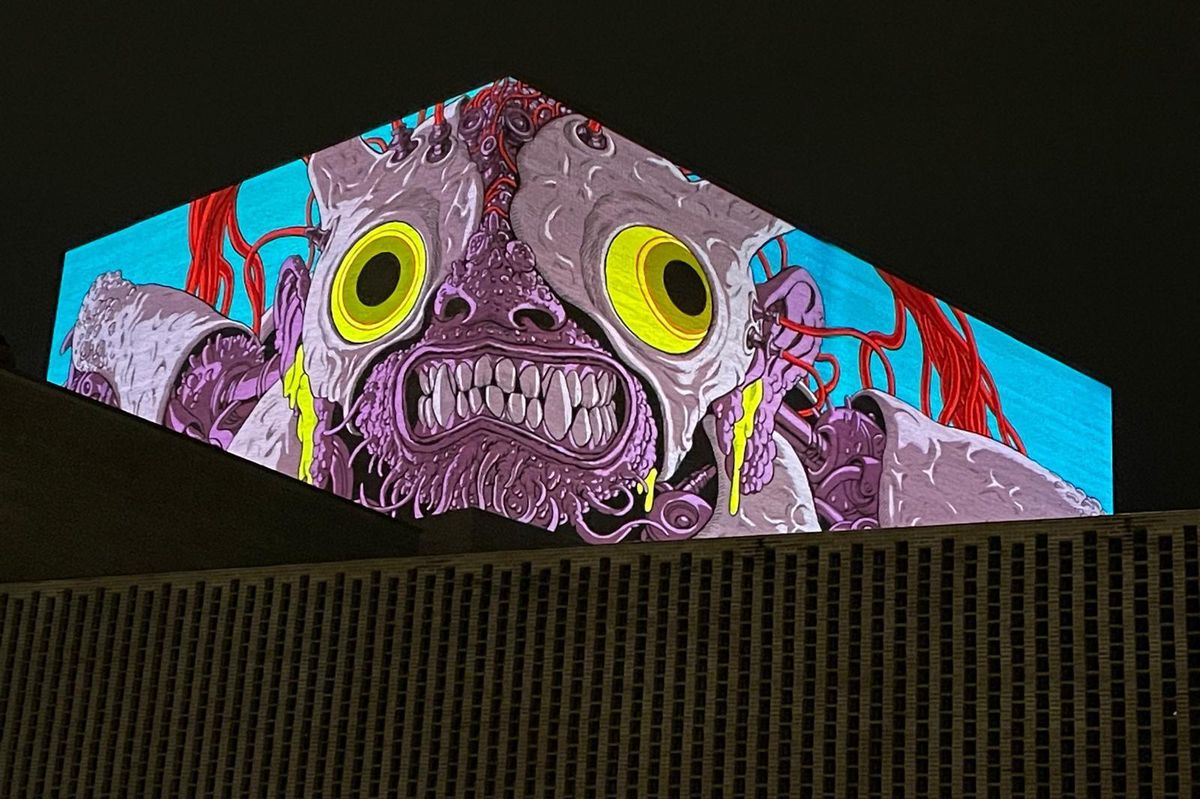 Projection d’une créature aux yeux jaunes et à la peau violette sur un bâtiment, créant une scène artistique saisissante avec un fond sombre.