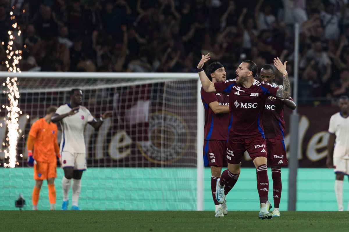 Des joueurs de football en tenue bordeaux célèbrent un but sur le terrain. Des étincelles jaillissent en arrière-plan.