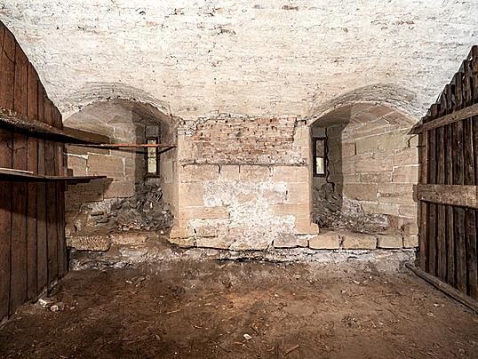 Intérieur d'une cave ancienne avec des murs en pierre, plafond voûté, sol en terre battue et étagères en bois.