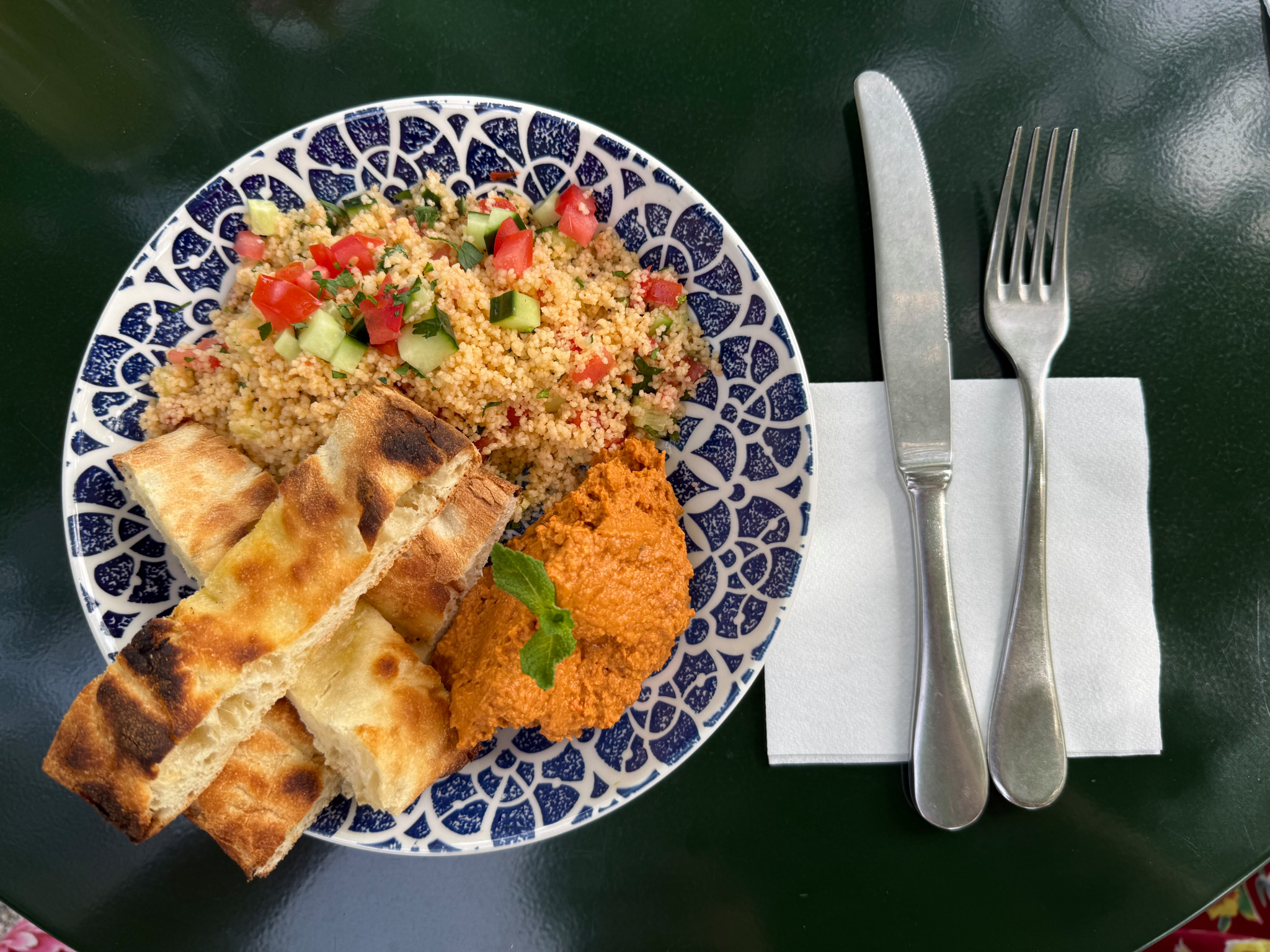 Teller mit Couscous-Salat, rotem Hummus und geröstetem Fladenbrot auf blau-weissem Teller, daneben Besteck und Serviette.